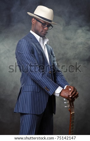 Black jazz musician wearing suit and white hat standing with guitar. Smoking cigarette and wearing glasses.