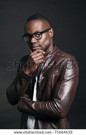 Confident casual young black man wearing brown leather jacket and glasses. Studio portrait against dark background.