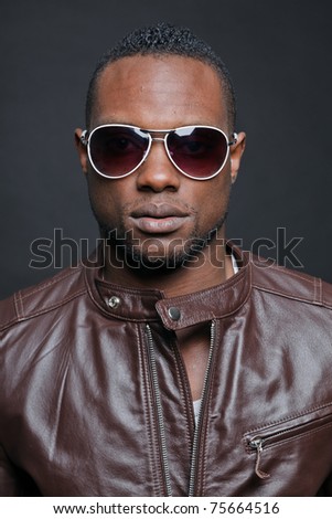 Confident casual young black man wearing brown leather jacket and sunglasses. Studio portrait against dark background.