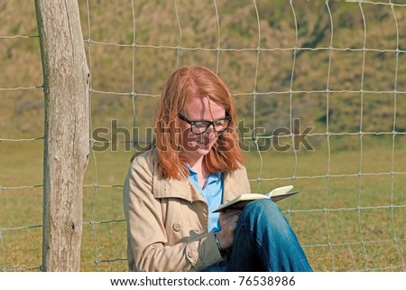 Young woman with red hair and glasses sitting in nature reading book.