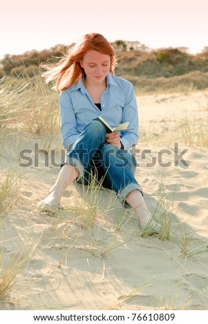 Casual young woman with red hair sitting in nature reading book.