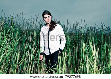 Young man with long brown hair standing in field with long grass. Stormy cloudy sky.