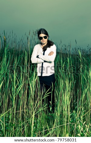 Young man with sunglasses and long brown hair standing in field with long grass. Stormy cloudy sky.