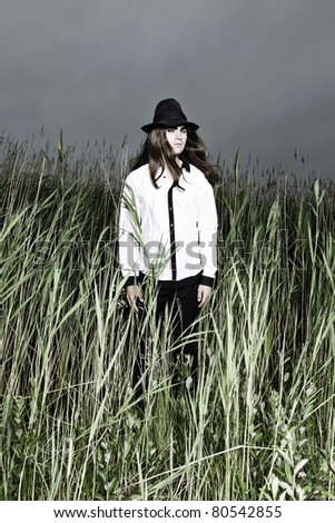 Young man with long brown hair wearing black hat standing in field with long grass. Stormy cloudy sky.