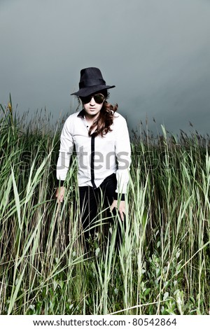 Young man with long brown hair wearing black sunglasses and black hat standing in field with long grass. Stormy cloudy sky.