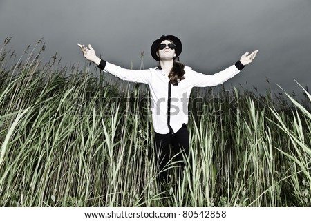 Young man with long brown hair wearing black sunglasses and black hat standing in field with long grass. Stormy cloudy sky.