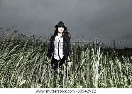 Young man with long brown hair wearing black suit and black hat standing in field with long grass. Stormy cloudy sky.
