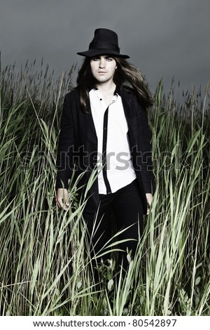 Young man with long brown hair wearing black suit and black hat standing in field with long grass. Stormy cloudy sky.