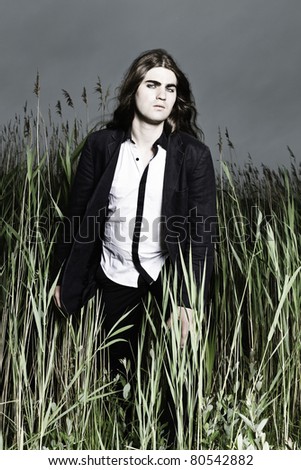 Young man with long brown hair wearing black suit standing in field with long grass. Stormy cloudy sky.