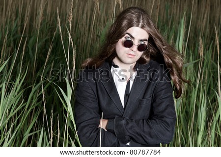 Young man with long brown hair wearing sunglasses and black suit standing in field with long grass. Stormy cloudy sky.