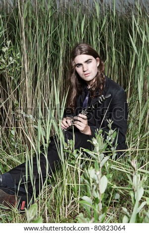 Young man with long brown hair wearing black suit holding clarinet in field of long grass. Stormy cloudy sky.
