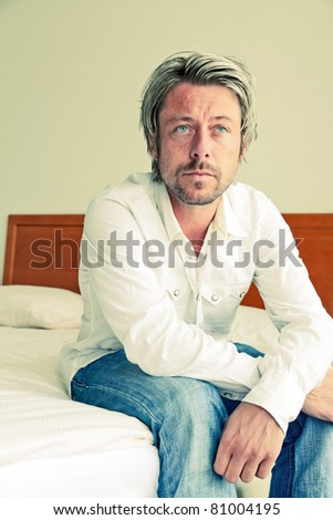 Young man with blond hair wearing white shirt sitting on bed in hotel room.