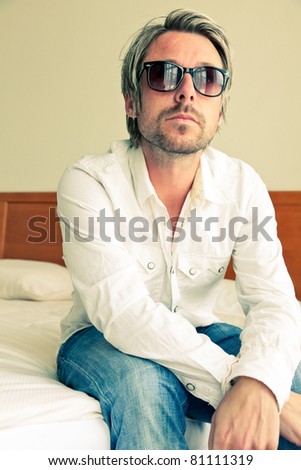 Young man with blond hair wearing sunglasses and white shirt sitting on bed in hotel room.
