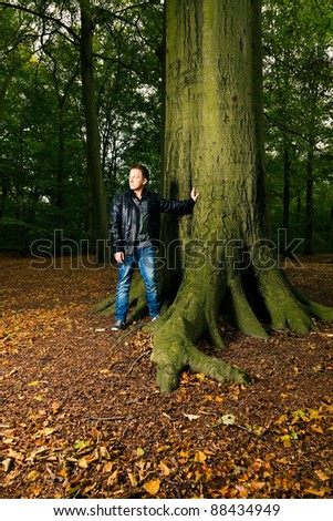 Single casual young man in forest leaning against tree. Short hair wearing jeans and black leather jacket.