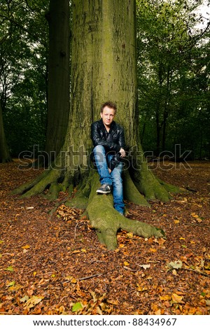 Single casual young man in forest sitting on tree. Short hair wearing jeans and black leather jacket.