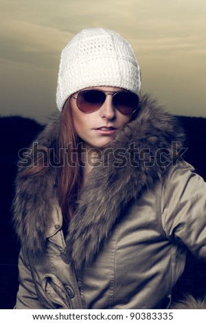 Close-up portrait of pretty young lady with red hair and blue eyes wearing white woolen cap and sunglasses and winter jacket. Outdoor at the beach in autumn.