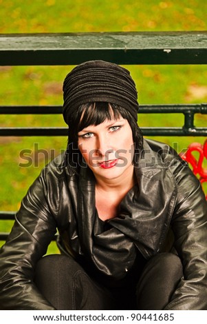 Pretty young woman with black leather jacket and black woolen cap sits in front of green fence. Grass and autumn leafs in the background.