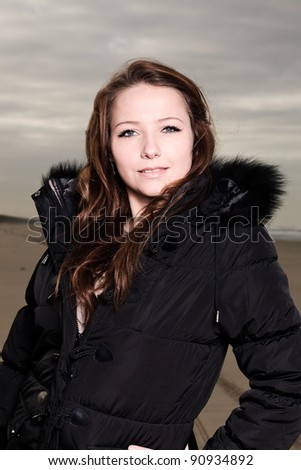 Pretty young woman with long brown hair wearing black winter coat on the beach. Autumn. Cloudy sky.