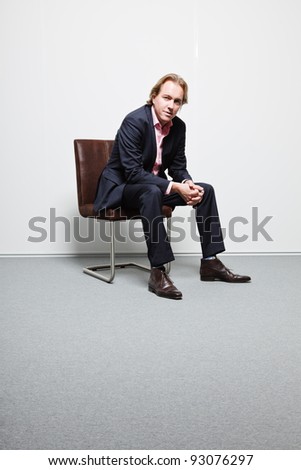 Young business man with blond hair in blue suit and pink shirt sitting on chair in office