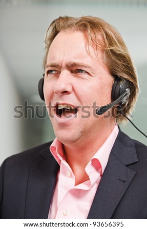 Young business man blond hair with headset wearing blue suit and pink shirt in front of window.