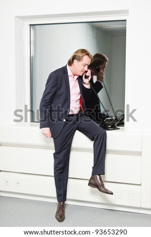 Young business man blond hair on the phone wearing blue suit and pink shirt. Standing in front of window.
