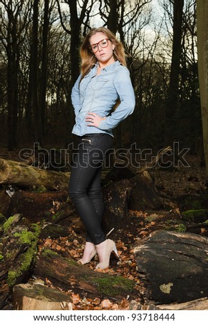Pretty young woman with long blond hair wearing glasses in winter forest. Wearing blue jeans shirt and black pants.