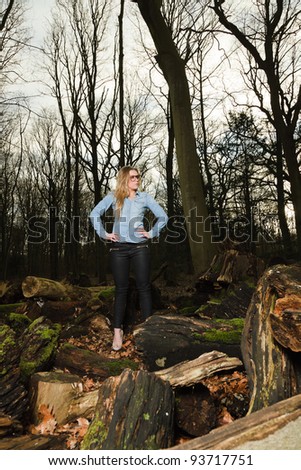 Pretty young woman long blond hair wearing glasses in winter forest. Wearing blue jeans shirt and black pants.