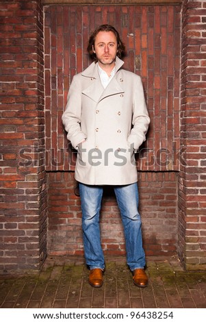 Young man with long hair wearing jeans and white jacket standing in front of brick wall