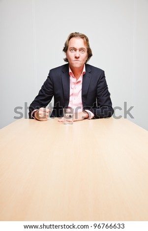 Angry business man with blond hair with glass of water sitting behind table in office isolated on white background