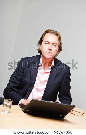 Business man with blond hair working with laptop computer sitting behind desk in office isolated on white background. Wearing pink shirt and blue suit.