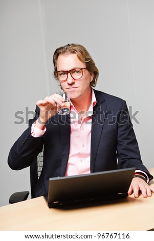 Business man with blond hair working with laptop computer sitting behind desk in office isolated on white background. Wearing pink shirt and blue suit.