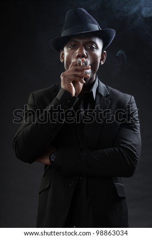 Young black man wearing suit and hat gangster style smoking cigar isolated on dark background. Studio portrait.