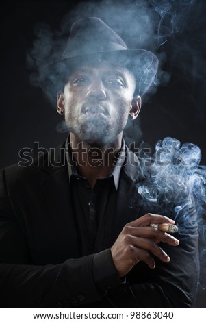 Young black man wearing suit and hat gangster style smoking cigar isolated on dark background. Studio portrait.
