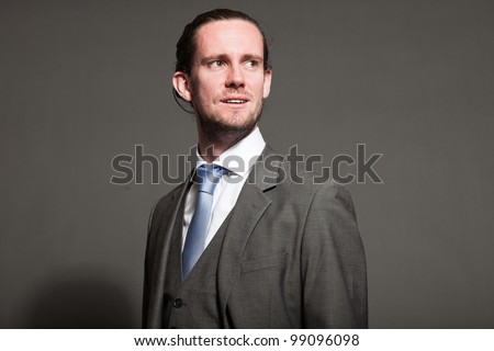 Young man brown long hair with expressive face wearing grey suit and blue tie. Isolated on grey background.