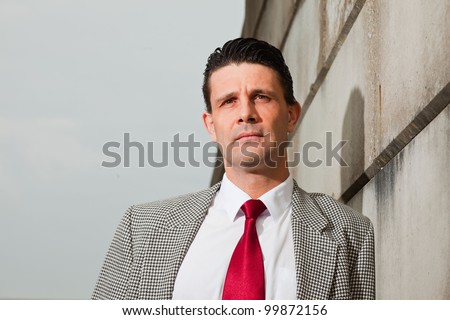 Business man with grey suit and red tie on the street leaning against brick wall. Industrial environment.