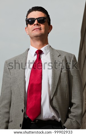 Business man with black vintage sunglasses and light grey suit and red tie walking on the street near brick wall. Industrial environment.