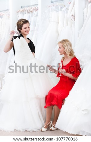 Shop assistant proposes another dress to the bride while she is drinking champagne