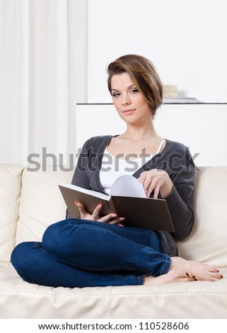 Pretty woman reads the book sitting on the white leather sofa
