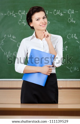 Smiley teacher at the blackboard holds 	blue writing pad
