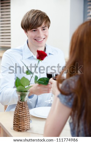 Pair is at the restaurant sitting at the table with vase and scarlet rose in it