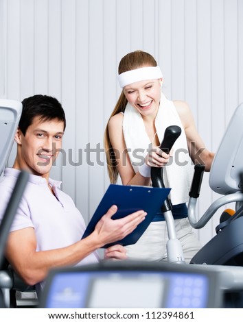 Young athletic woman training on gym equipment in gym with coach writing down the results