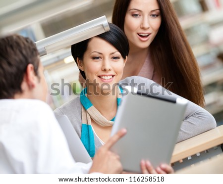 Man sitting at the table at the library shows something interesting in the pad to two women