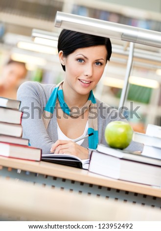 Woman with green apple surrounded with piles of books reads sitting at the table at the library. Learning process