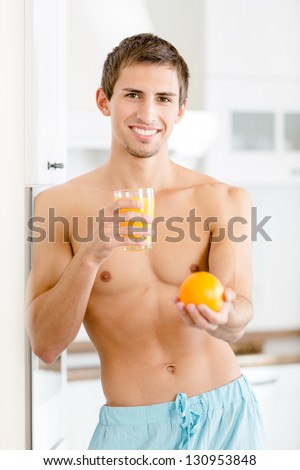 Half-naked man with glass of juice and orange standing near the fridge at the kitchen