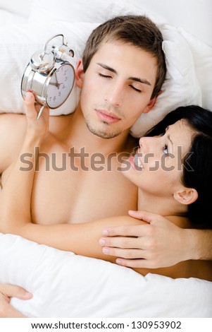 Close up of couple lying in bedroom. Woman holds alarm clock near the ear of man, top view