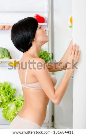 Woman seeks food in the opened refrigerator full of vegetables and fruit. Concept of healthy and dieting food