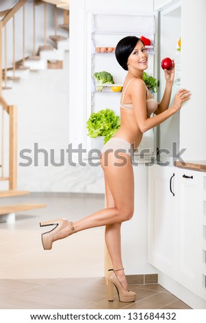 Full-length portrait of woman near the opened refrigerator full of vegetables and fruit. Concept of healthy and dieting food