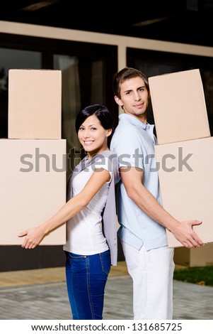 Happy couple stands back to back while carrying cardboard boxes and moving to new house