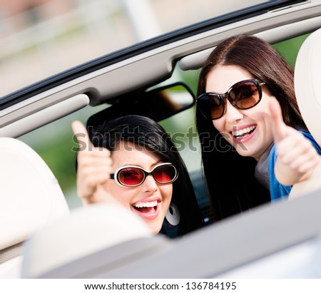 Two happy girls sitting in the car and thumbing up turn back and have fun while having little car trip