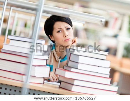Sad female student is tired of education surrounded with piles of books sitting at the table